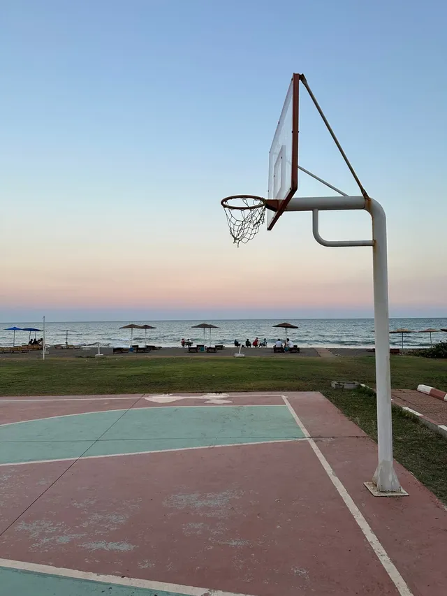 An outdoor basketball hoop on a seafront court at sunset, with parasols and the sea behind